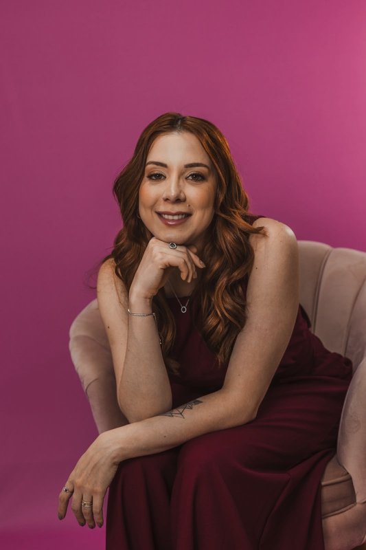Dra. Maria Luiza Garbiere sentada em poltrona clara, com a mão no queixo, sobre fundo magenta